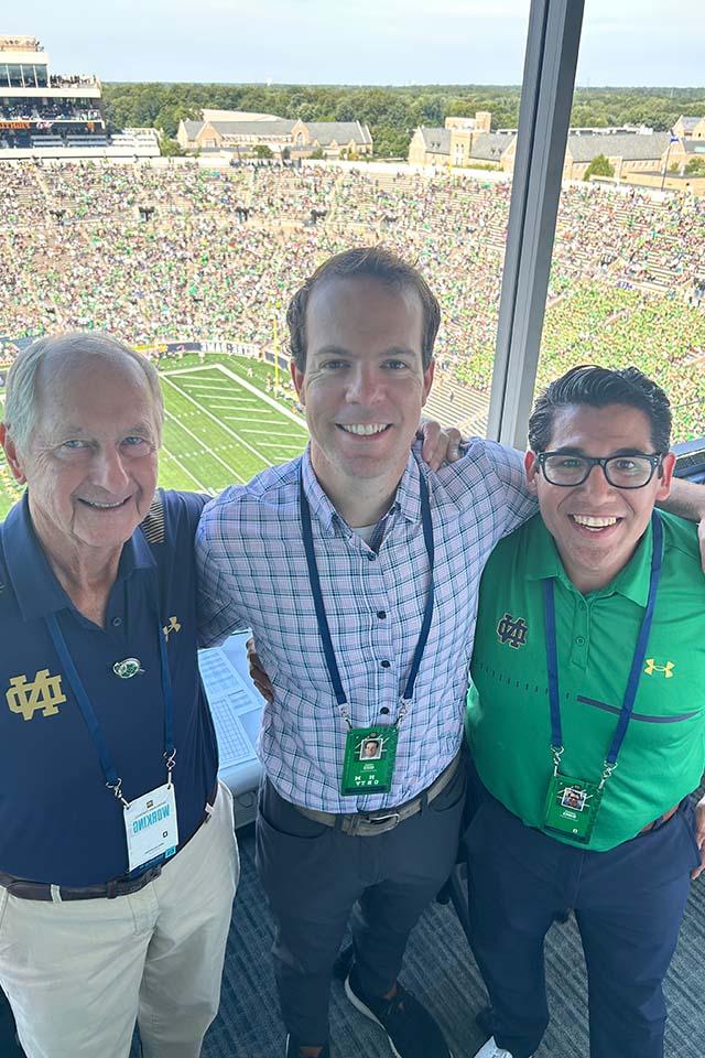 Three men pose for a photo with a stadium behind them. Two of the men are wearing Notre Dame shirts.